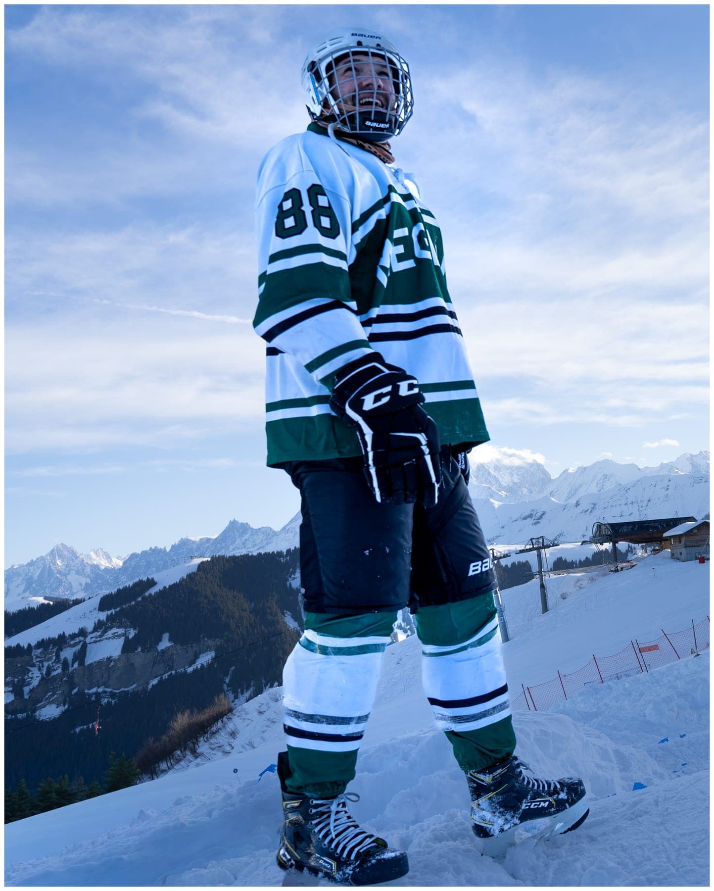 A rider in a green-and-white hockey jersey standing on the slope, mountains behind