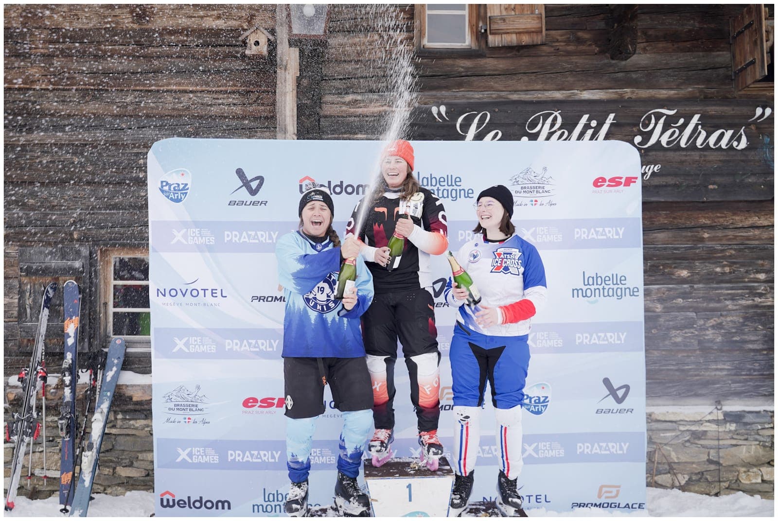 Three riders on the podium spraying champagne in front of a wooden chalet