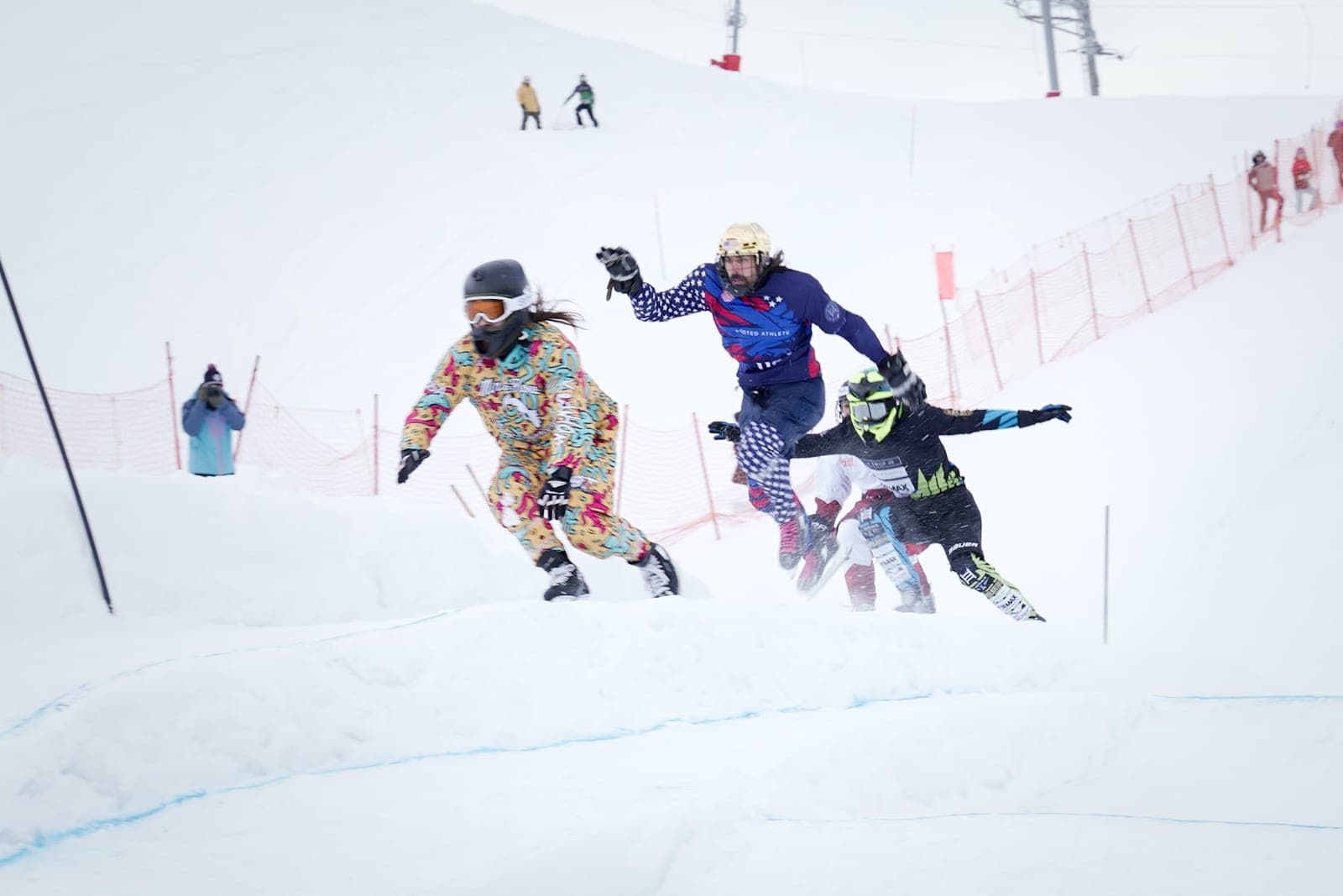Four riders bunched over a bump on a snowy track, one in a patterned suit in the lead